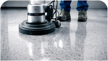 Floor buffer/polisher cleaning a speckled concrete or terrazzo floor, reflecting light as it polishes the surface.