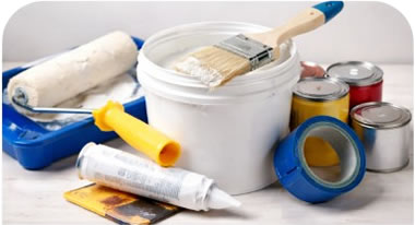 Assorted painting supplies on a worktable: a white paint bucket with brush, rollers, tape, and paint cans.