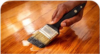 Hand holding a wide flat brush applying wood finish to a polished hardwood floor.