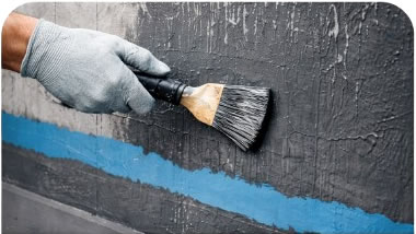 Gloved hand painting a concrete wall with dark paint over a blue stripe.